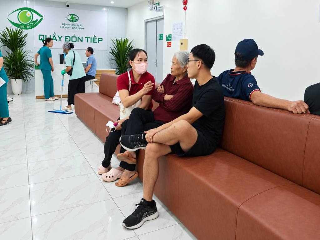 Những người ngồi gần quầy đón tiếp; phía sau có nhân viên y tế và công việc làm sạch sàn.