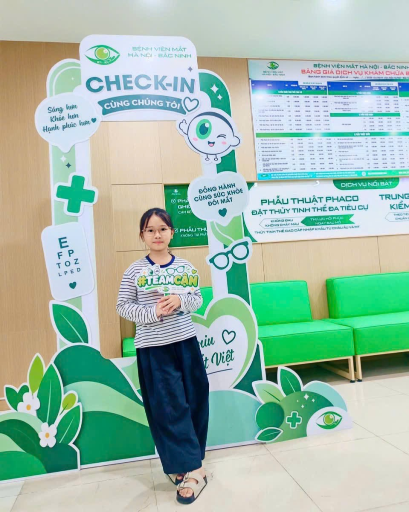 Khách nhỏ tuổi đeo kính đứng tại khung check-in Bệnh viện Mắt Hà Nội – Bắc Ninh.