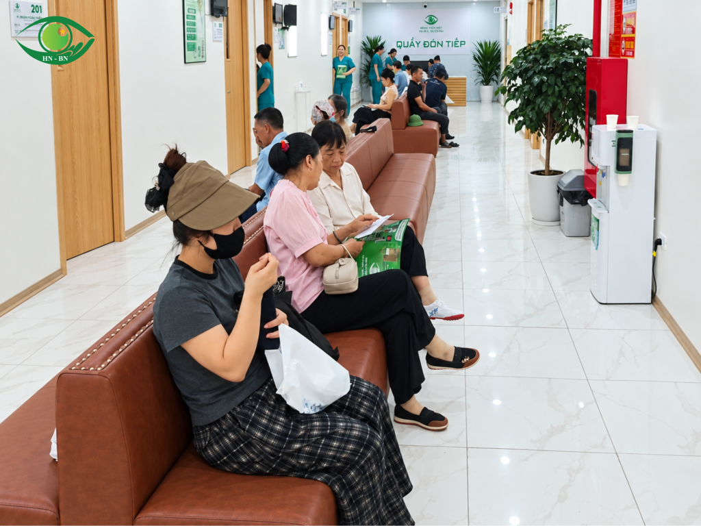 Nhiều người ngồi chờ dọc hành lang các phòng khám, một số người cầm giấy tờ hoặc tài liệu.