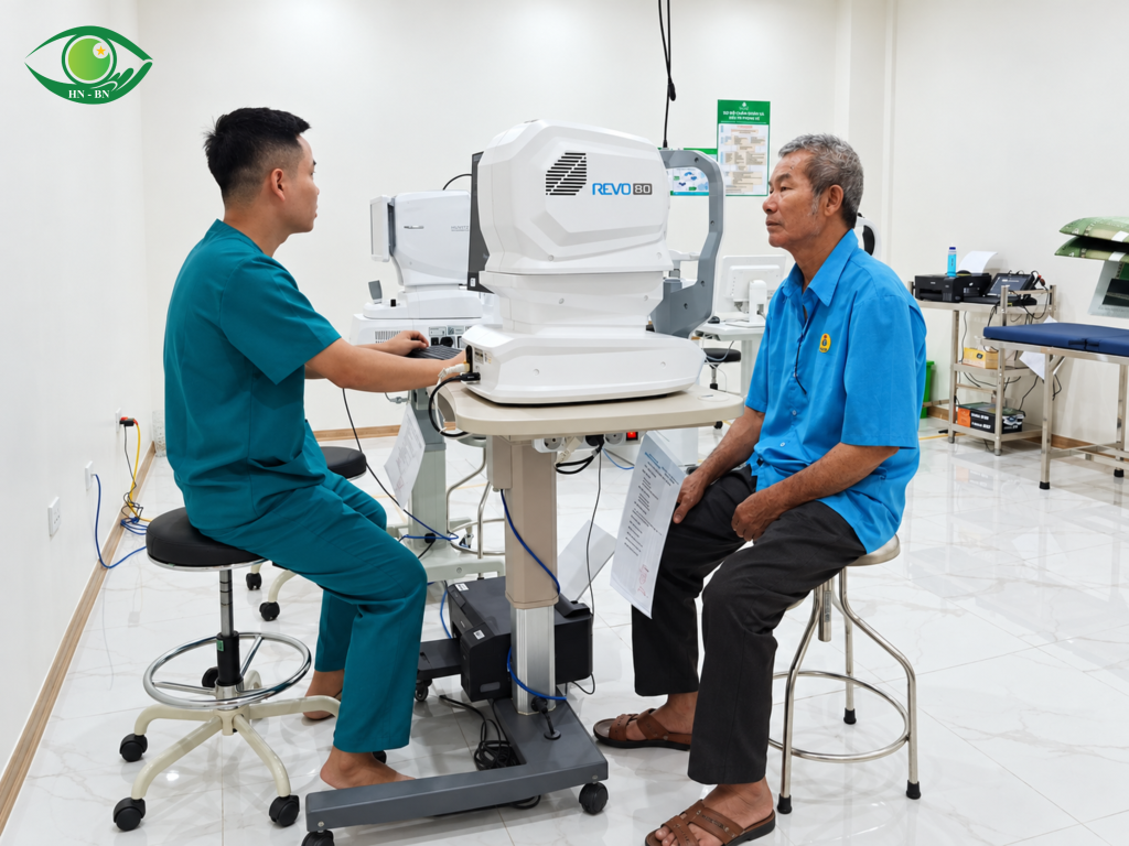 Người đàn ông ngồi đối diện thiết bị nhãn khoa, nhân viên y tế ở bên bàn điều khiển.