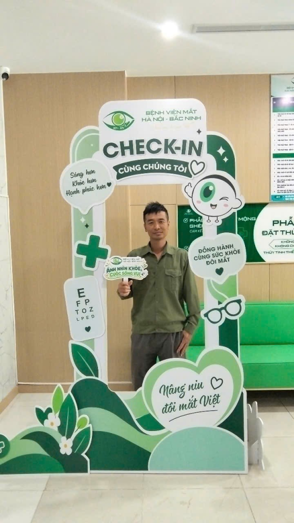 Người đàn ông đứng trong khung check-in màu xanh, cầm bảng “Ánh nhìn khỏe, cuộc sống vui”.