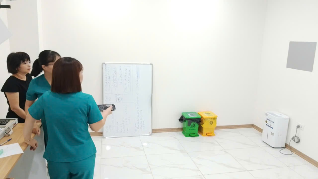 Nhân viên y tế cầm thiết bị điều khiển trong phòng kiểm tra, bên cạnh là một người phụ nữ và một nhân viên khác.