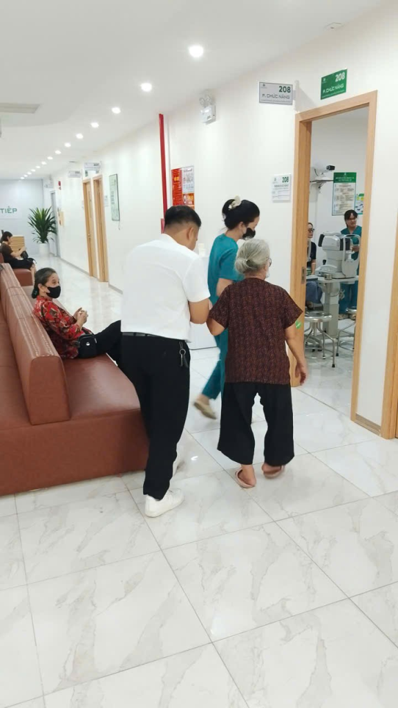 Người phụ nữ lớn tuổi được hỗ trợ di chuyển về phía phòng chức năng, nhân viên y tế đi phía trước.