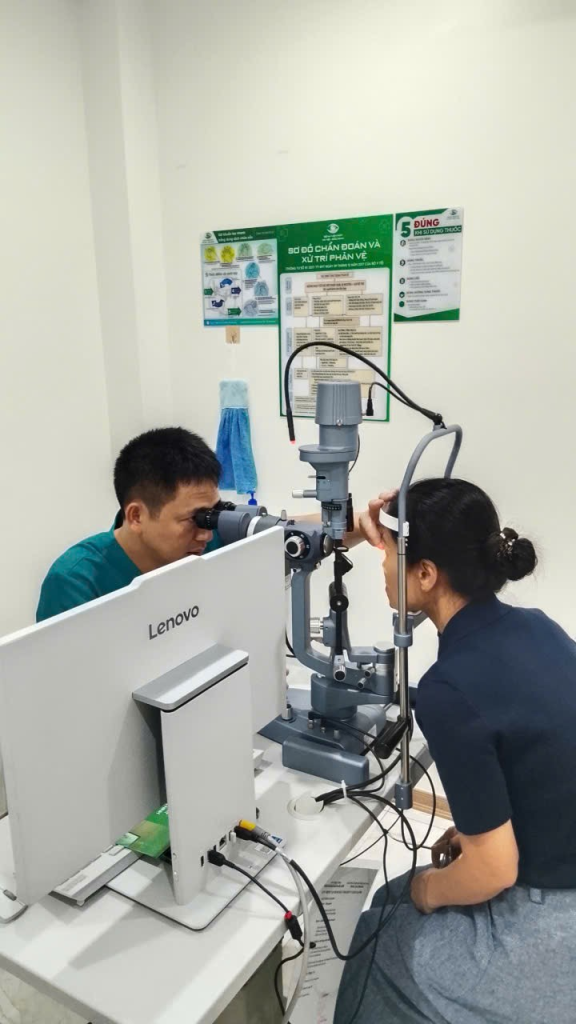 Người phụ nữ ngồi trước thiết bị khám mắt, nhân viên y tế quan sát và hỗ trợ vùng mắt.