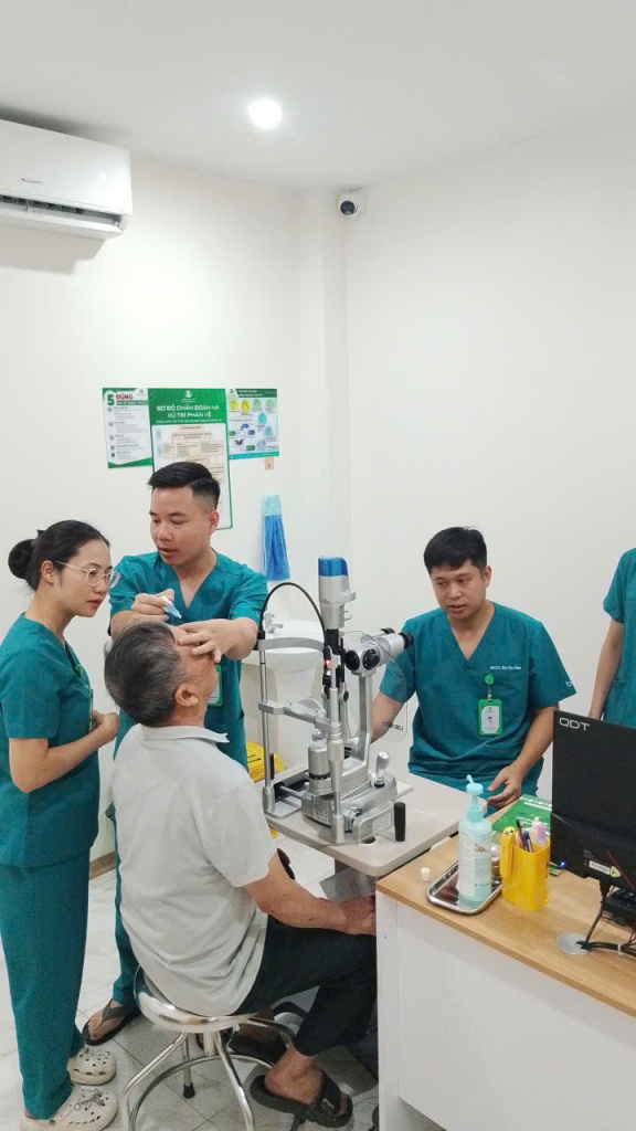Người bệnh lớn tuổi ngồi bên thiết bị khám mắt, nhiều nhân viên y tế phối hợp hỗ trợ.